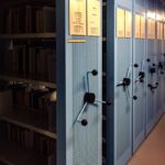 Rows of blue archival cabinets with open shelves displaying various documents and files in a dimly lit storage area